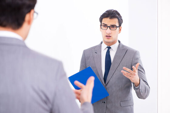 Man in grey suit with glasses holding blue folder talking in mirror