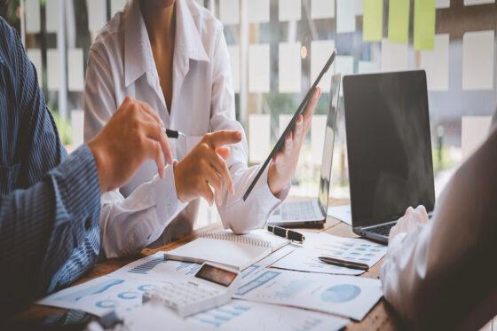 Sales reps gathered around a table reviewing data on tablets, computers, and paper